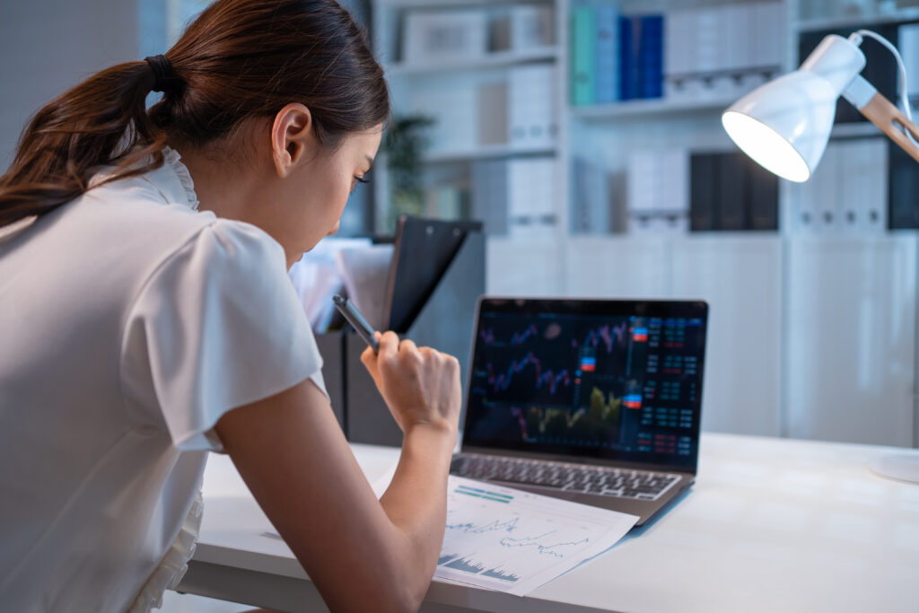 An Asian woman wearing a white shirt with her hair in a ponytail sits at a white table holding a pen in her right hand. In front of her is a printed image of some financial charts and a laptop with live financial charts is open in front of her. In the background there are white shelves and a white lamp.