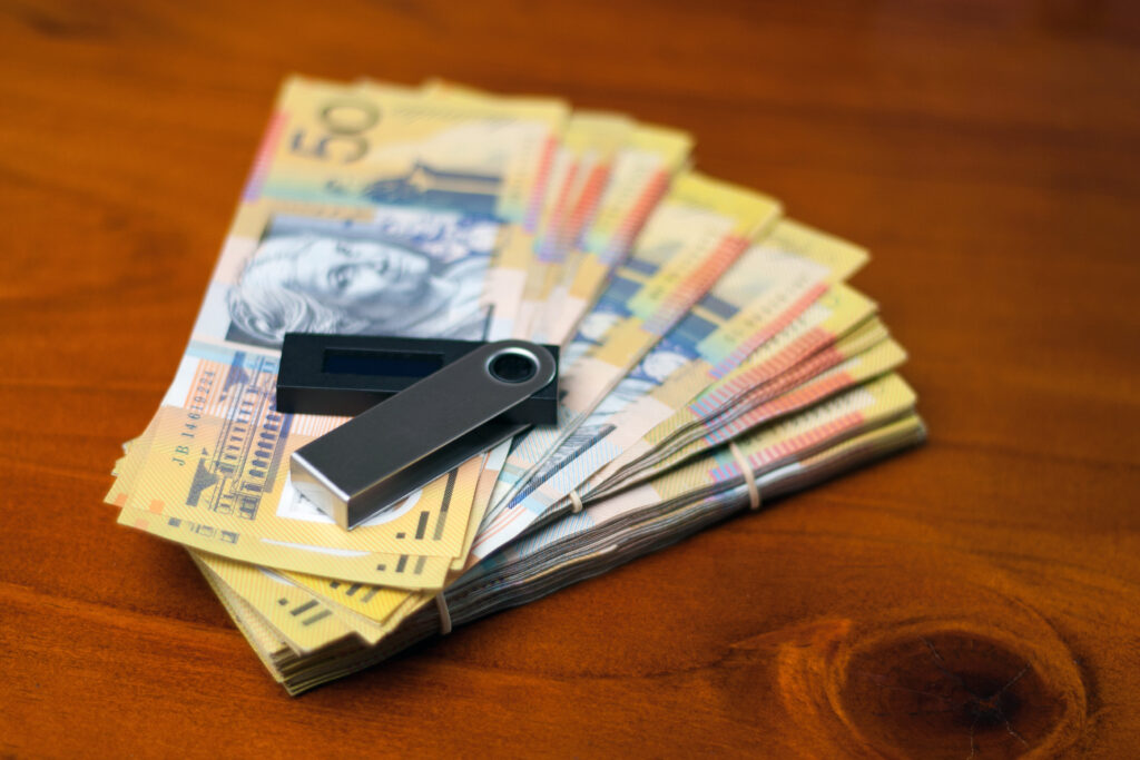 A Ledger Nano X sitting on top of a pile of Australian fifty-dollar bills on top of a wooden surface.