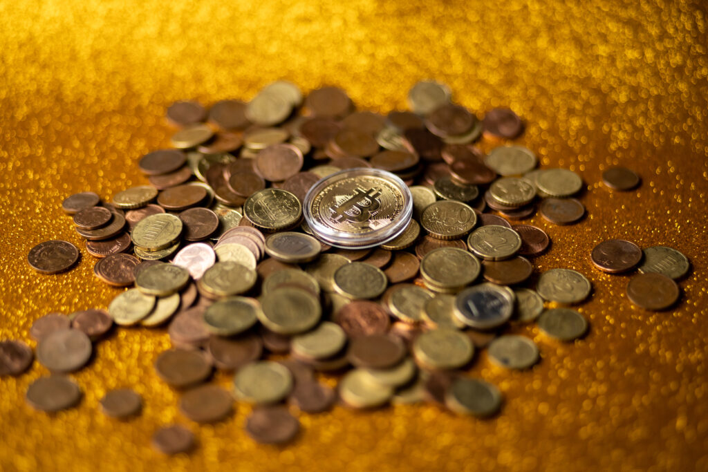 A pile of gold and bronze coins on a shimmery dark gold surface. On the top of the pile of coins is a large gold coin with the Bitcoin logo emblazoned on it.