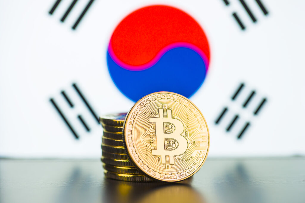 A stack of golden coins with the bitcoin logo embossed on it, in front of the South Korean flag.