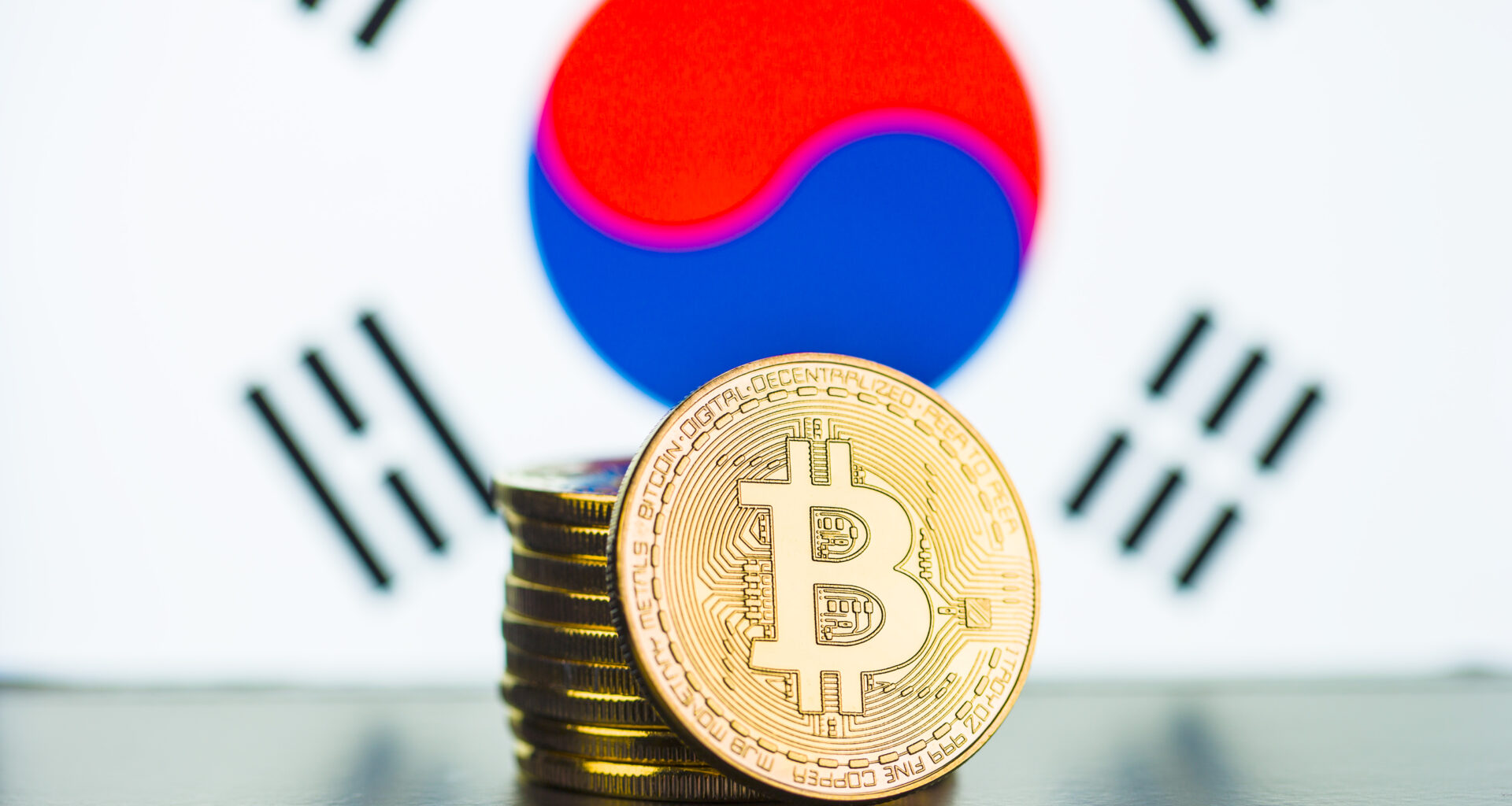 A stack of golden coins with the bitcoin logo embossed on it, in front of the South Korean flag.