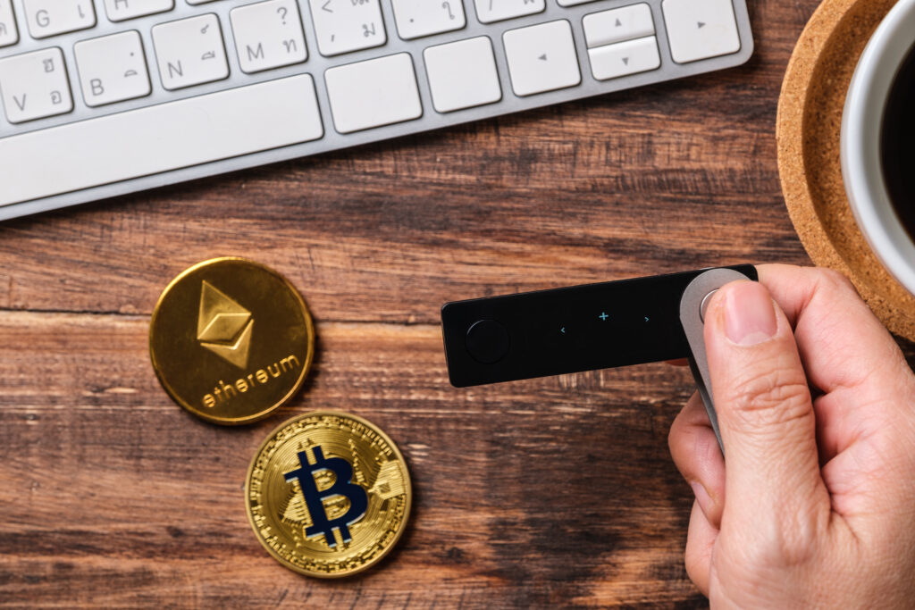 A hand is holding a crypto hardware wallet, and there are two gold coins beside it. One of them is an ETH gold coin, and the other is a gold Bitcoin.