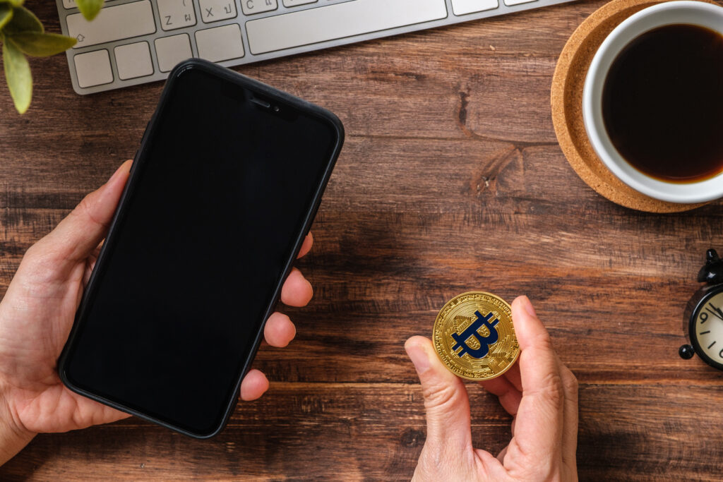 A left hand holding a phone with a dark screen on the left, and a right hand holding a gold coin with the Bitcoin logo on the right. The upper right corner has a cup of coffee, and the bottom of a keyboard is visible near the top of the image. 
