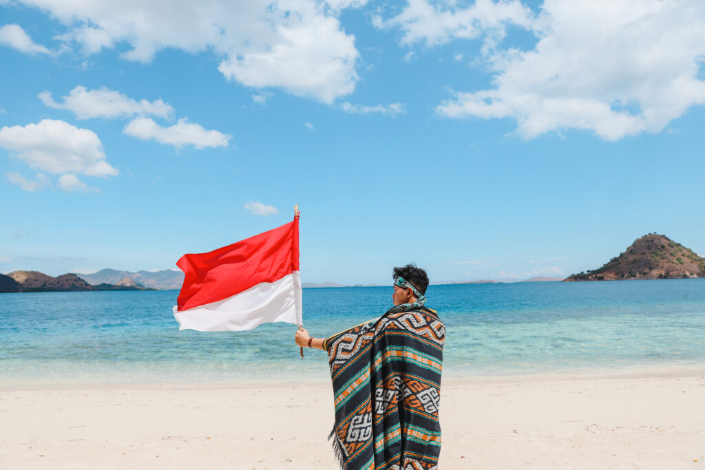 A man is holding up an Indonesian flag in his left hand. He is standing on a beach with blue sea and sky.
