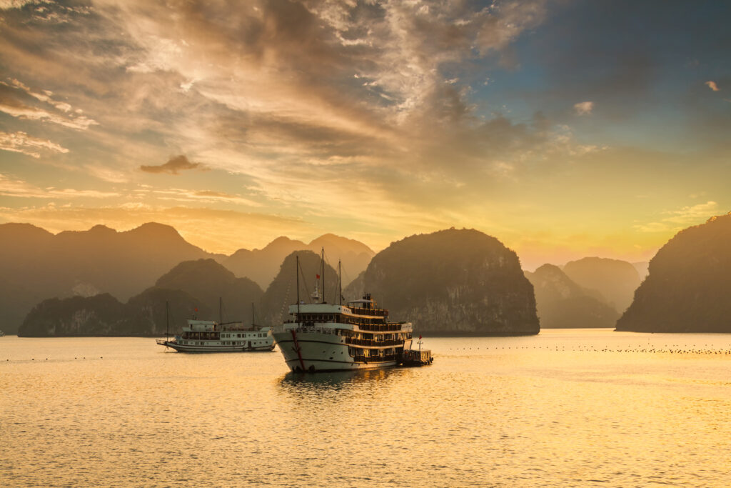 Sunset over the islands of Halong Bay in Vietnam. There are two ships in the foreground and a golden sky as the backdrop.