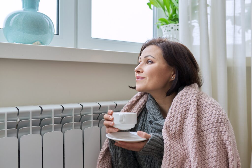 Winter season, woman warming up near home heating radiator. Female in warm woolen sweater under blanket with cup of hot drink