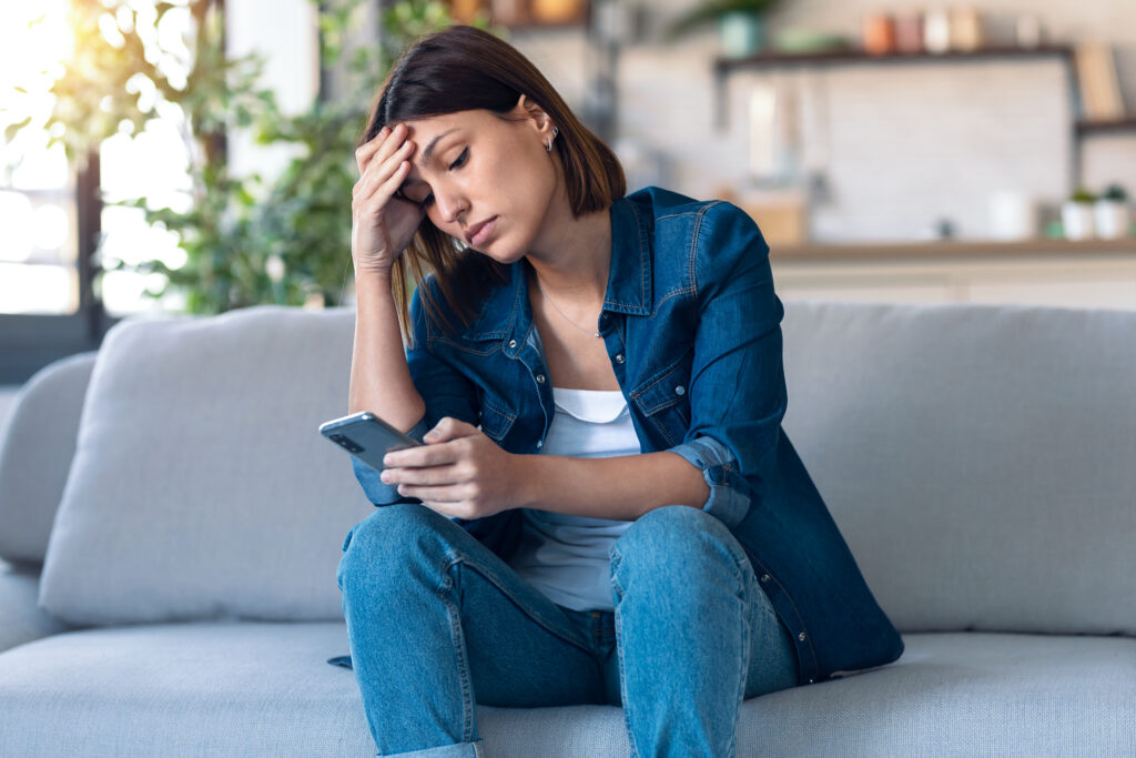 Shot of worried young woman using her mobile phone while thinking about problems sitting on couch in the living room at home.