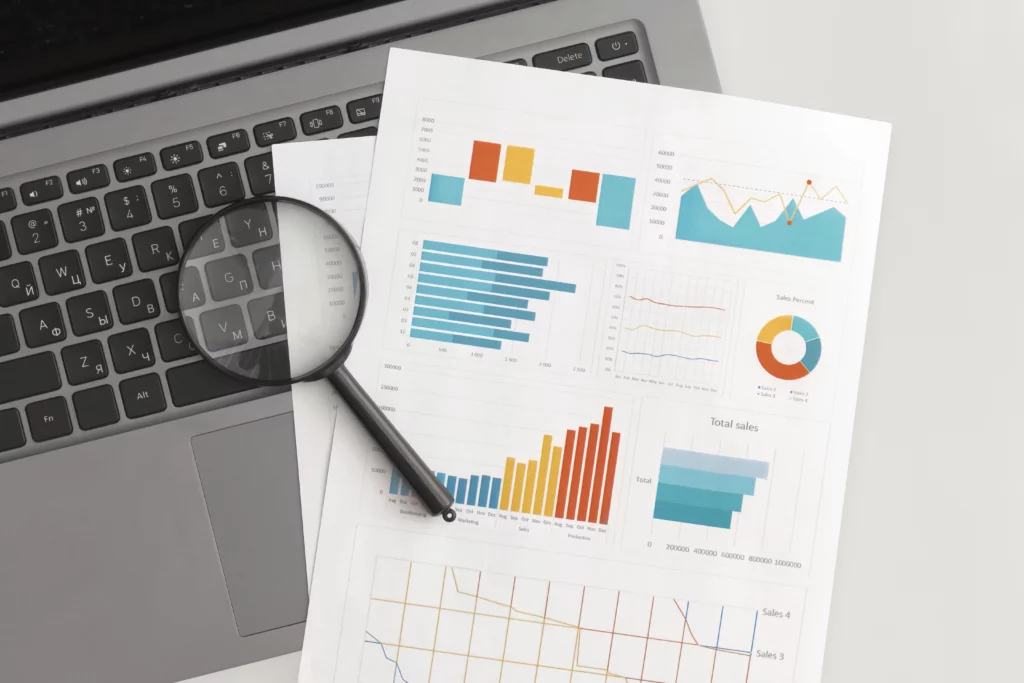 a magnifying glass sits on top of a paper depicting various types of charts and an open laptop