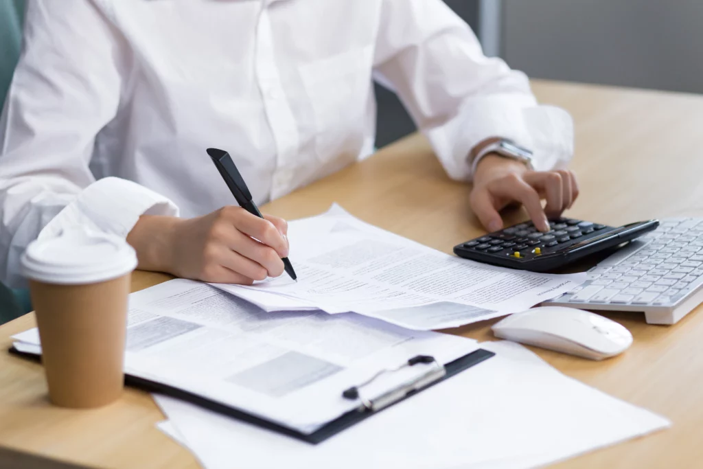 a person wearing a white shirt is tapping on a calculator and writing something at the same time, with a cup of coffee at their side. their face cannot be seen.