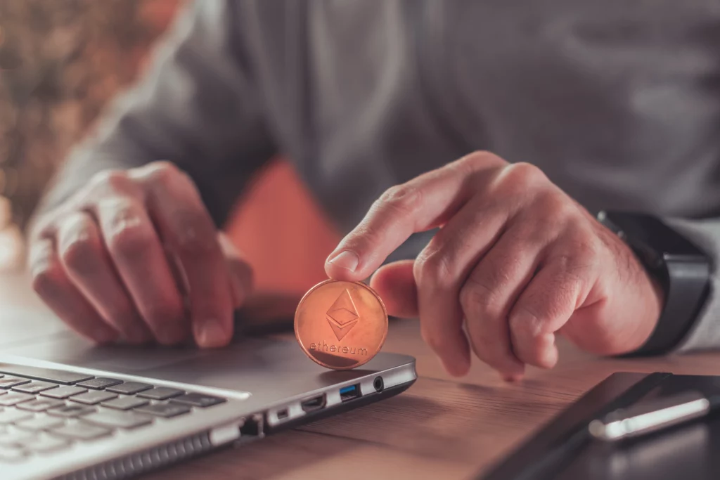 A man in a long sleeved business shirt is sitting at a desk with one hand on his laptop and another hand holding an Ether coin that he is showing to the camera.