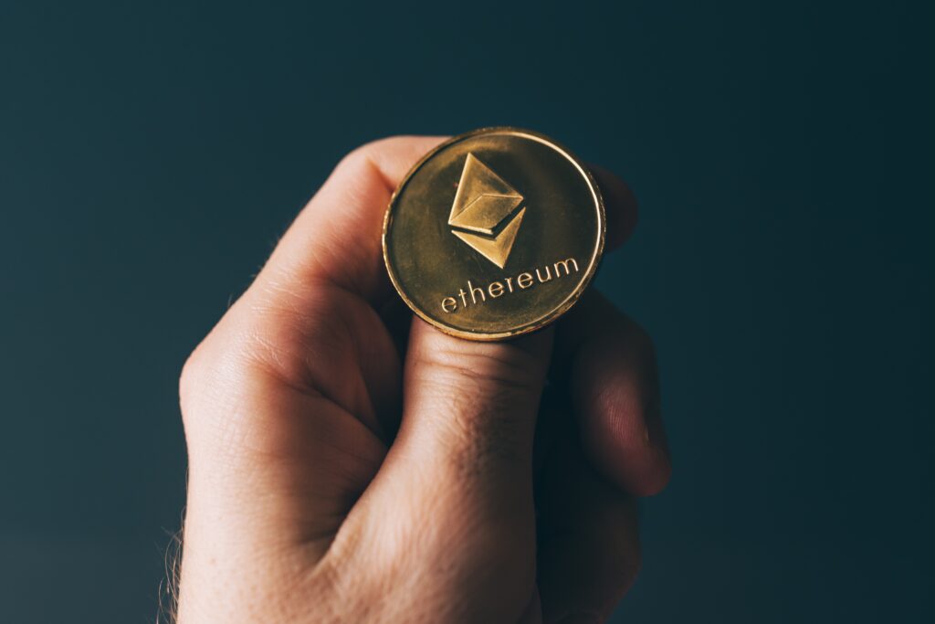 A hand has a gold Ethereum coin balanced on the thumb as though it's about to flip it.