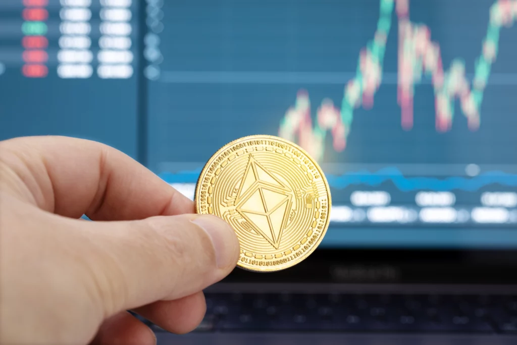 A hand is holding a gold Ether coin in front of a financial chart.