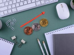 Three Bitcoin coins are lined up on an office desk with stationery around it. Above the coins is a red arrow that is pointing diagonally upwards to signify economic growth.