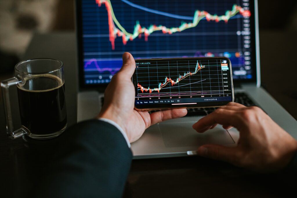 A man is holding a phone in front of a laptop. Both screens are showing economic graphs.