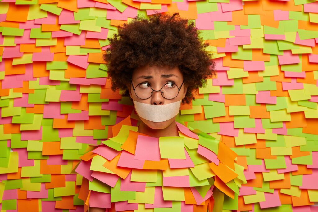 A woman is standing in front of a colourful background with tape across her mouth and a confused look on her face.