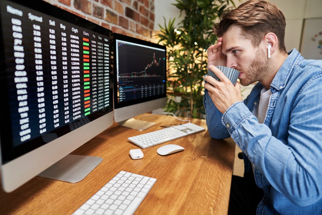 A white man in a blue shirt sits on the right side of the image, drinking from a blue cup and looking at 2 computer screens filled with financial charts and information.