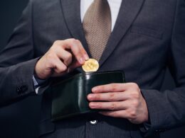 A man in a business suit is holding a wallet and a gold Bitcoin.