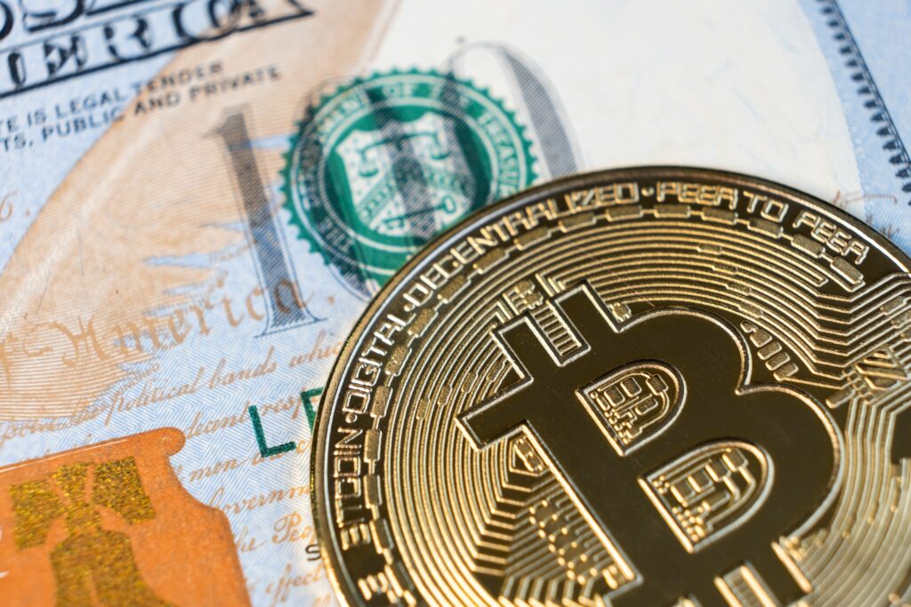 The image shows a gold embossed Bitcoin on top of an American hundred-dollar bill.