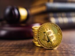 A stack of gold Bitcoins sit on a wooden table in front of a gavel.