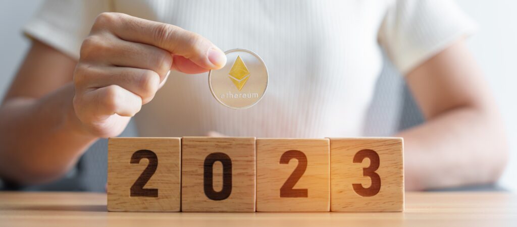 A person in a white shirt is holding up a gold Ether coin above blocks that say "2023".