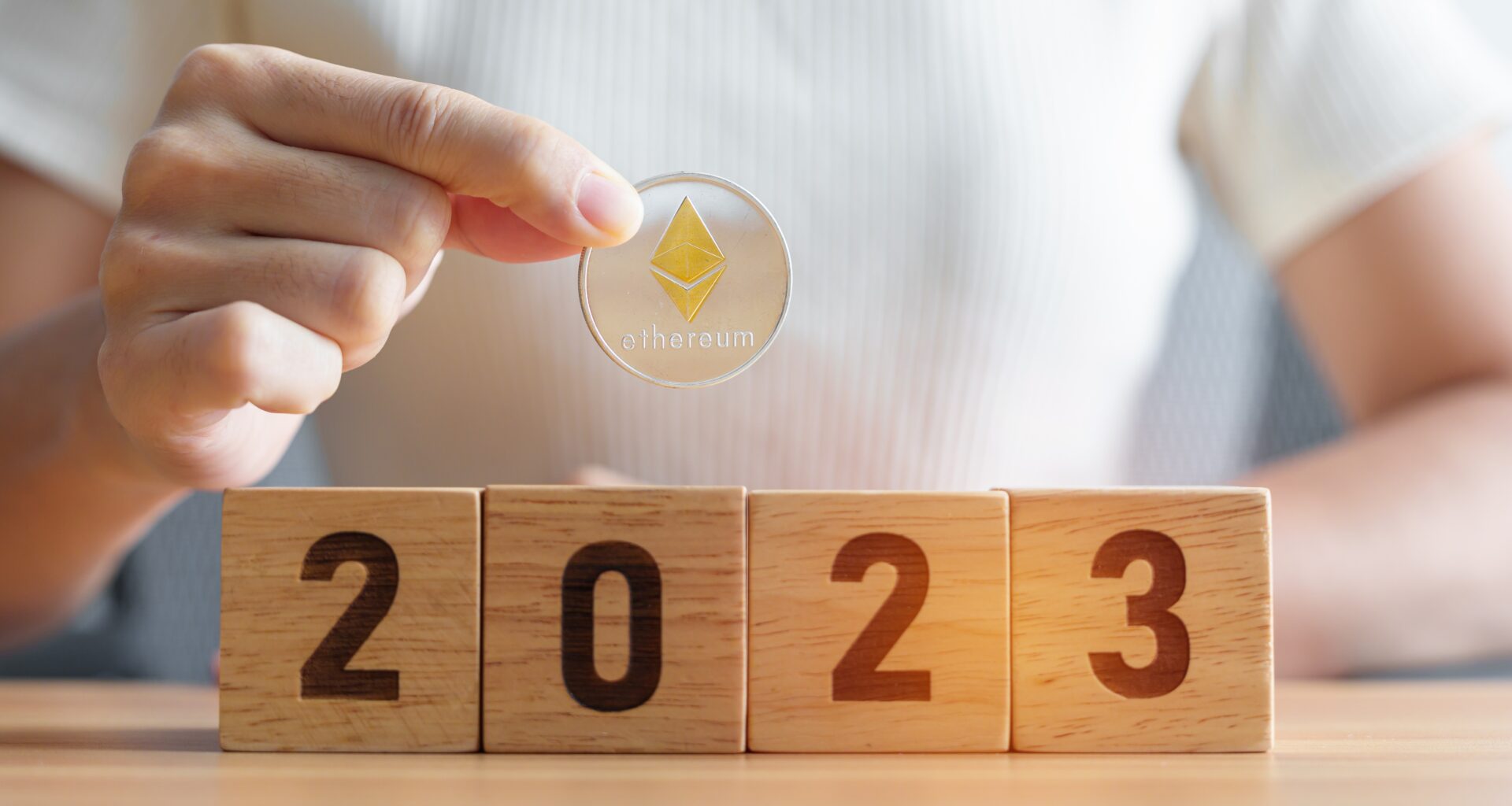 A person in a white shirt is holding up a gold Ether coin above blocks that say "2023".